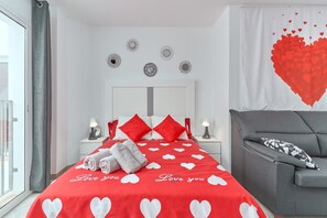 A bedroom with a red heart-patterned bedspread, a grey sofa, and a large red heart decoration on the wall.