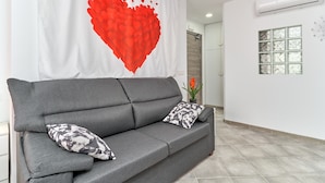 Un canapé gris agrémenté de coussins à motifs, un mur orné d’un cœur rouge et une fenêtre à vitres dépolies.