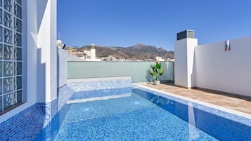A rooftop pool with blue mosaic tiles, a potted plant, and a view of mountains in the background.