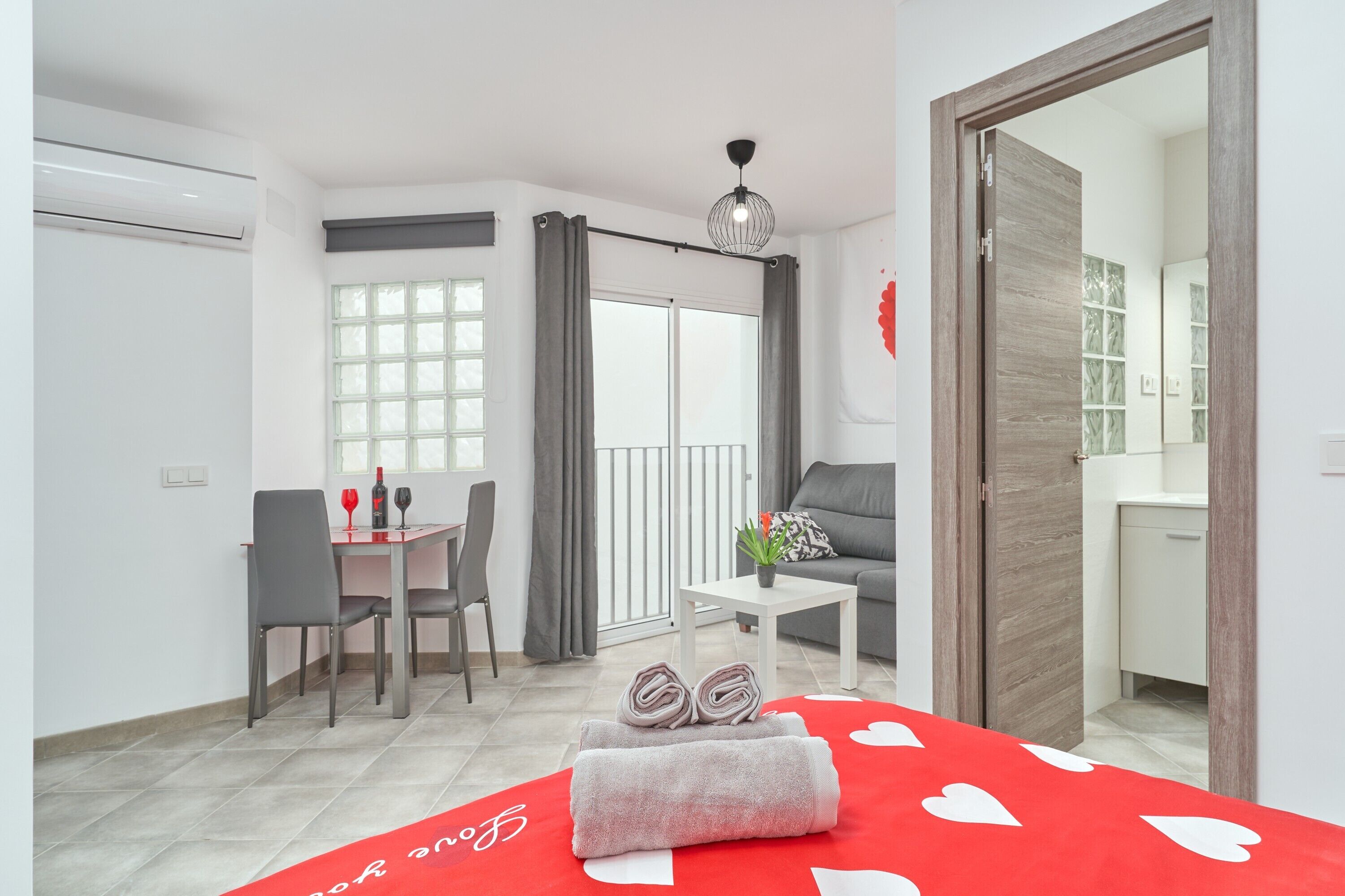 A hotel room with a dining area, a sofa, a small table with a plant, a bed with a red heart-patterned bedspread, and a sliding door leading to a bathroom.
