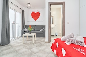 A modern bedroom with a grey sofa, a small table with a plant, a red bedspread with heart patterns, and a large red heart decoration on the wall.