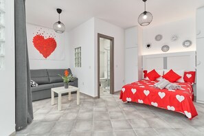 A modern bedroom with a grey sofa, a small table with a plant, a bed with red and white heart-patterned bedding, and a large red heart artwork on the wall. - Studio Apartment "Nerja Boutique Sweet Corner B3" with Shared Pool & Wi-Fi (Nerja)
