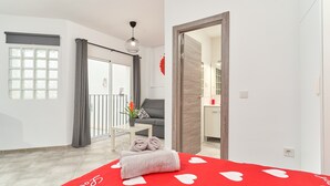 A hotel room with a bed, a small table with a plant, a grey sofa, and a balcony with a railing.
