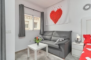A modern room with a grey sofa, a white table with a plant, a red heart-themed wall hanging, and a window with grey curtains. - Studio Apartment "Nerja Boutique Deluxe 103" with Shared Pool & Wi-Fi (Nerja)