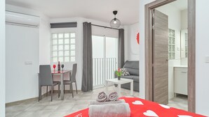 Une chambre d’hôtel comprenant un coin repas, un canapé, une petite table avec une plante, un lit recouvert d’une courtepointe à motifs de cœurs rouges, et une porte coulissante donnant accès à la salle de bain.