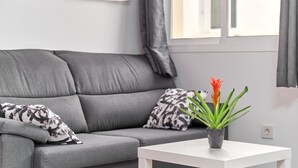 Un canapé gris agrémenté de coussins à motifs, une table d’appoint blanche et une plante en pot aux feuilles vertes et à la fleur orange.