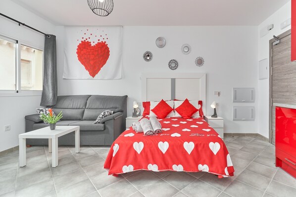 Une chambre à coucher avec un lit, un canapé gris, une table blanche et une couvre-lit aux motifs de cœurs rouges.