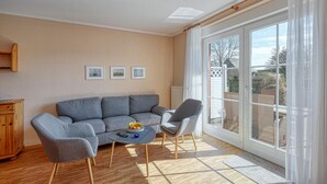 Una sala de estar con un sofá gris, un sillón gris y una pequeña mesa redonda con un frutero. En la pared, tres cuadros enmarcados, un aparador de madera y un amplio ventanal corredero que da a un espacio exterior.
