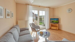 Una sala de estar con un sofá, una silla, una mesita con un frutero, un televisor sobre un mueble de madera y una ventana con cortinas.