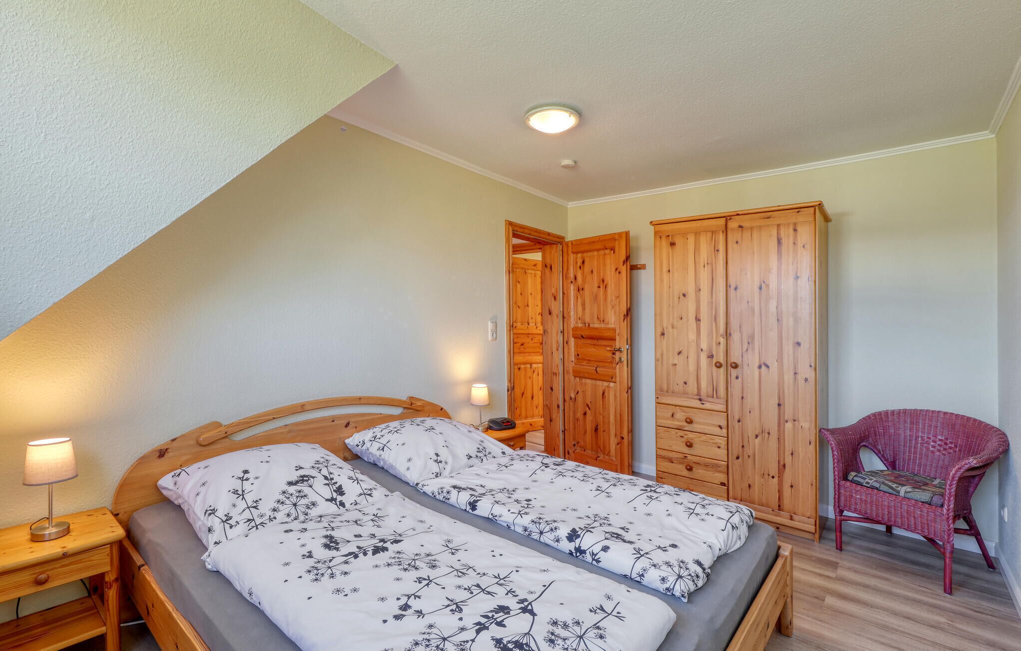 Un dormitorio con una cama de madera, una mesita de noche con lámpara, un sillón de mimbre rojo y un armario de madera.