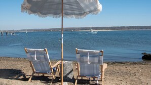 Dos sillas de playa bajo una sombrilla blanca en una playa arenosa, con vista a barcos en el agua.