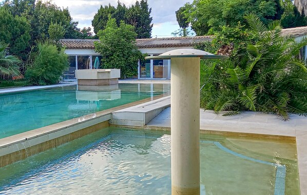 A swimming pool with a stone structure in the center, surrounded by lush greenery and a building with a tiled roof. - Lovely home in Aramon (Aramon)