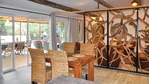 Een eetgedeelte met rieten stoelen en een houten tafel, een glazen scheiding met een houten sculptuur, en uitzicht op de omgeving door grote ramen.