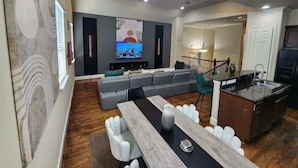 A modern living room with a grey sectional sofa, a dining table with white chairs, and a kitchen area with a sink and stove. - Horizon House | Dual Home |Private Pool |Sleeps 16 (Houston)