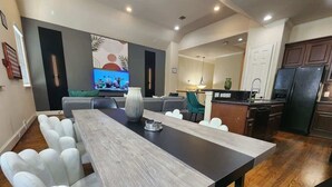 A modern dining area with a long table, white chairs, and a television in the background. - Horizon House | Dual Home |Private Pool |Sleeps 16 (Houston)