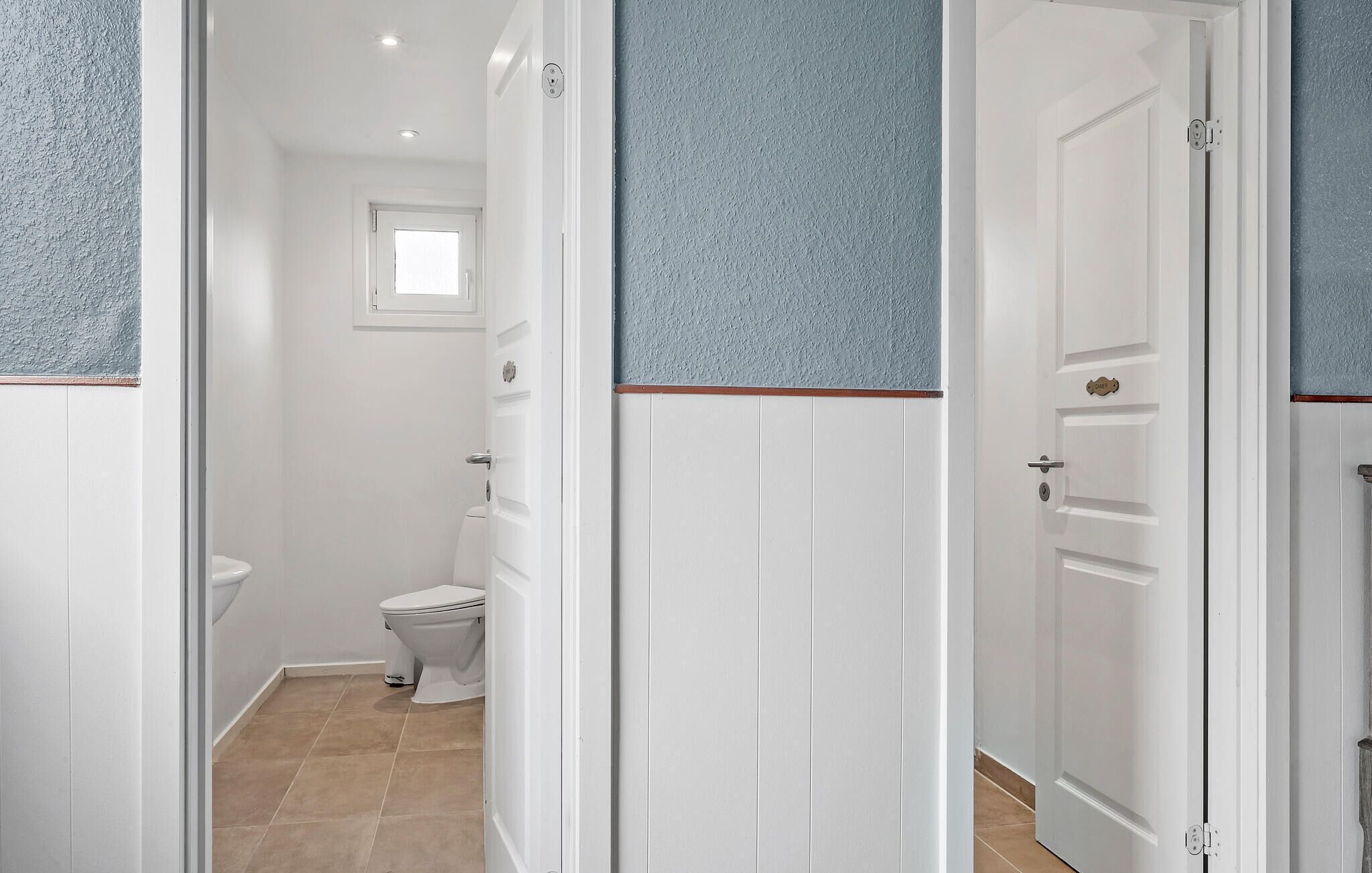 A narrow hallway with white doors and blue textured walls, leading to a bathroom with a toilet and a window.