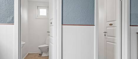 A narrow hallway with white doors and blue textured walls, leading to a bathroom with a toilet and a window.