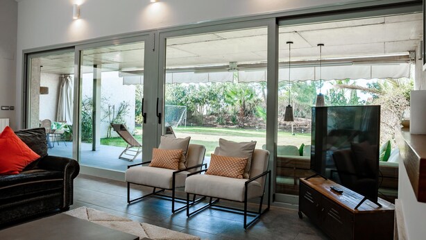 Een moderne woonkamer met een glazen schuifdeur naar een achtertuin, twee fauteuils met kussens, een donker lederen bank, een houten kast en een televisie.
