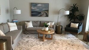 A living room with a grey sofa, a round coffee table with a plant, a painting of a beach, and a tall floor lamp. - Beautiful Home on a canal.  Heated pool.  Sunrise and Sunsets are spectacular. (Cape Coral)