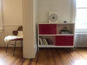 Una habitación con suelo de madera, una estantería blanca y roja, una silla blanca con un cojín blanco mullido y un radiador.