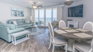A modern living room with a sofa, coffee table, and dining table. There is a ceiling fan, a TV mounted on the wall, and a view of the ocean through large windows. - BIG DISCOUNTS ON ALL MONTHLY RENTALS  - EXLARGE FACING  Emerald Coast Gulf Front (Panama City Beach)