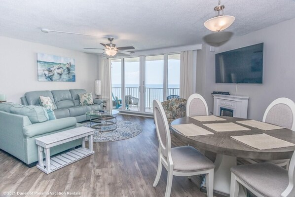 A modern living room with a sofa, coffee table, and dining table. There is a ceiling fan, a TV mounted on the wall, and a view of the ocean through large windows.