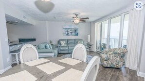 A spacious living room with a dining area, a ceiling fan, a large window with curtains, and a balcony with a chair. - BIG DISCOUNTS ON ALL MONTHLY RENTALS  - EXLARGE FACING  Emerald Coast Gulf Front (Panama City Beach)