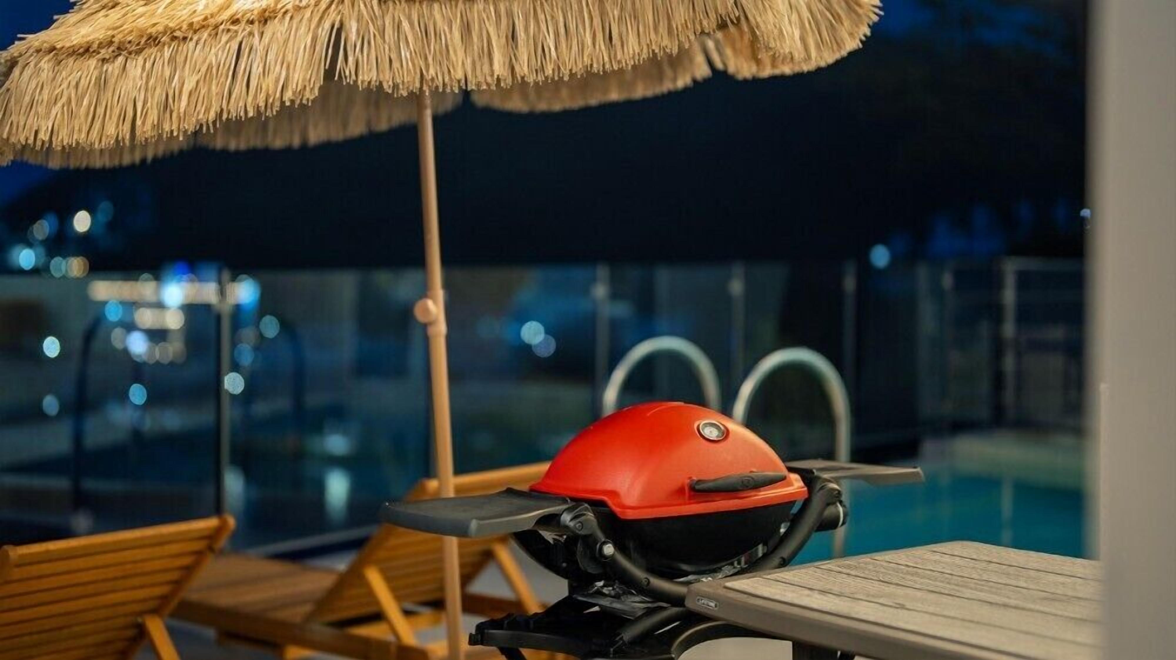 Un espace aménagé au bord de la piscine, avec un barbecue rouge, des chaises longues en bois et un parasol en chaume.