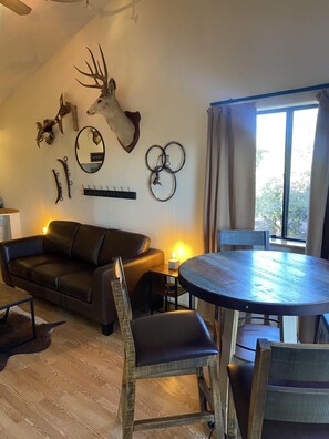 Un salon chaleureux avec un canapé en cuir, une table ronde en bois et un coin repas. Une tête de cerf est accrochée au mur, et divers éléments décoratifs ornent les murs, notamment un miroir et un porte-manteau.