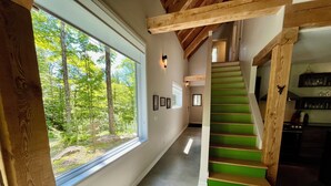 Un escalier en bois, avec des marches vertes, mène au deuxième étage. Une grande fenêtre sur le côté gauche offre une vue sur une forêt.