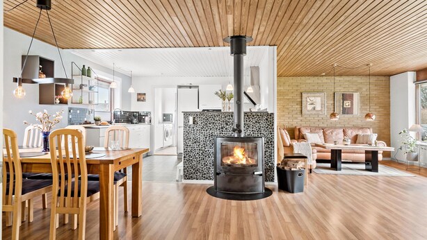 A modern living room with a central wood-burning stove, a dining area with wooden chairs, and a kitchen with white cabinets and a stone backsplash.