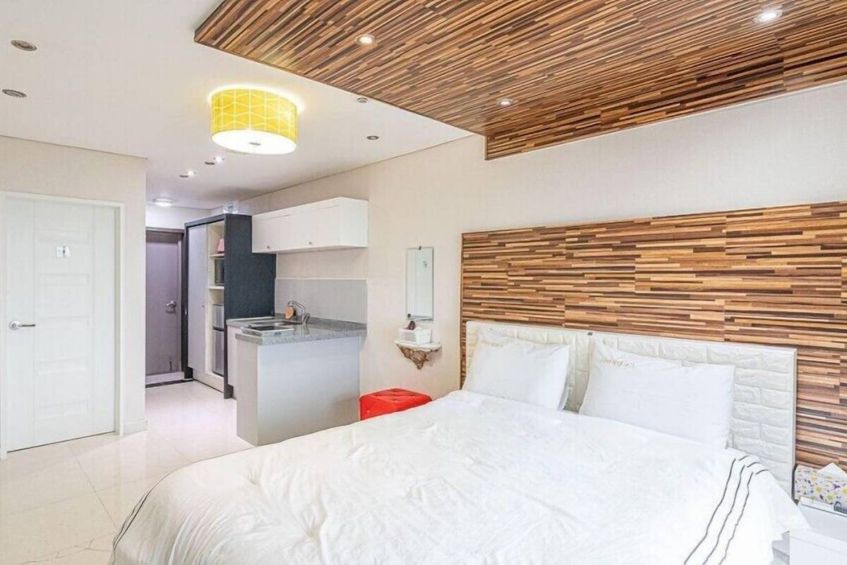 Habitación de hotel moderna con una cama grande, cabecera de madera, kitchenette pequeño y baño visible a través de una puerta abierta.