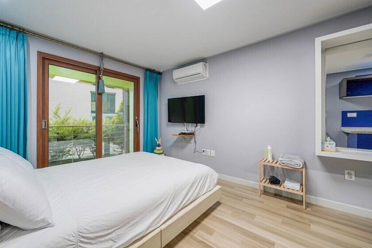 Un dormitorio con cama, televisor, un pequeño estante de madera y ventana con cortinas.