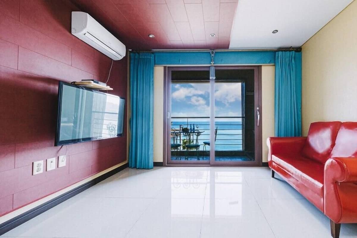 Una sala de estar moderna con un sofá rojo, un televisor de pantalla plana en la pared y un ventanal con vistas al mar.