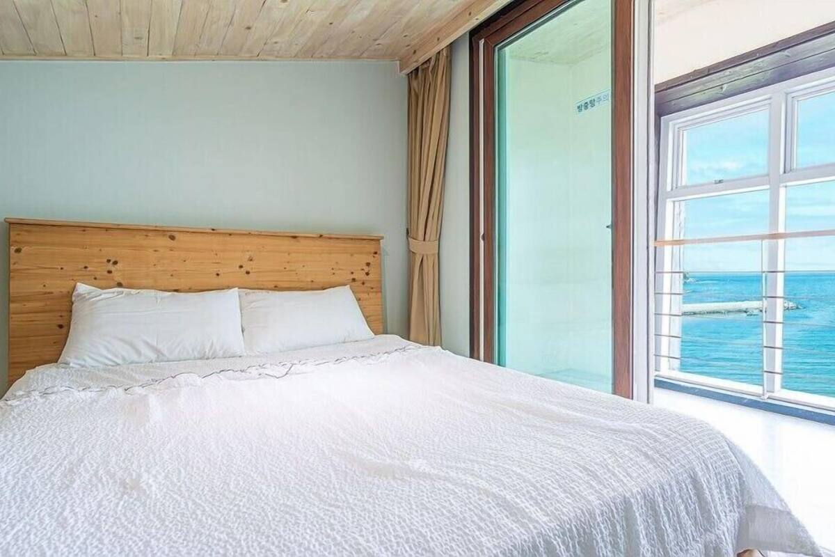 Un dormitorio con cabecera de madera, ropa de cama blanca y un ventanal con vista al mar.