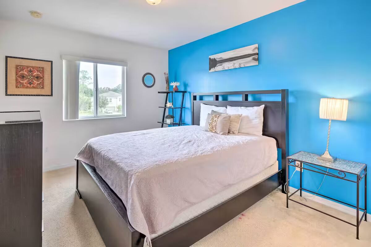 A bedroom with a bed, a nightstand, a lamp, a window, and a wall with a blue accent.