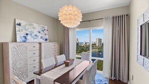 Uma sala de jantar com mesa de madeira, cadeiras brancas, um lustre decorativo e vista para a paisagem urbana através de grandes janelas.