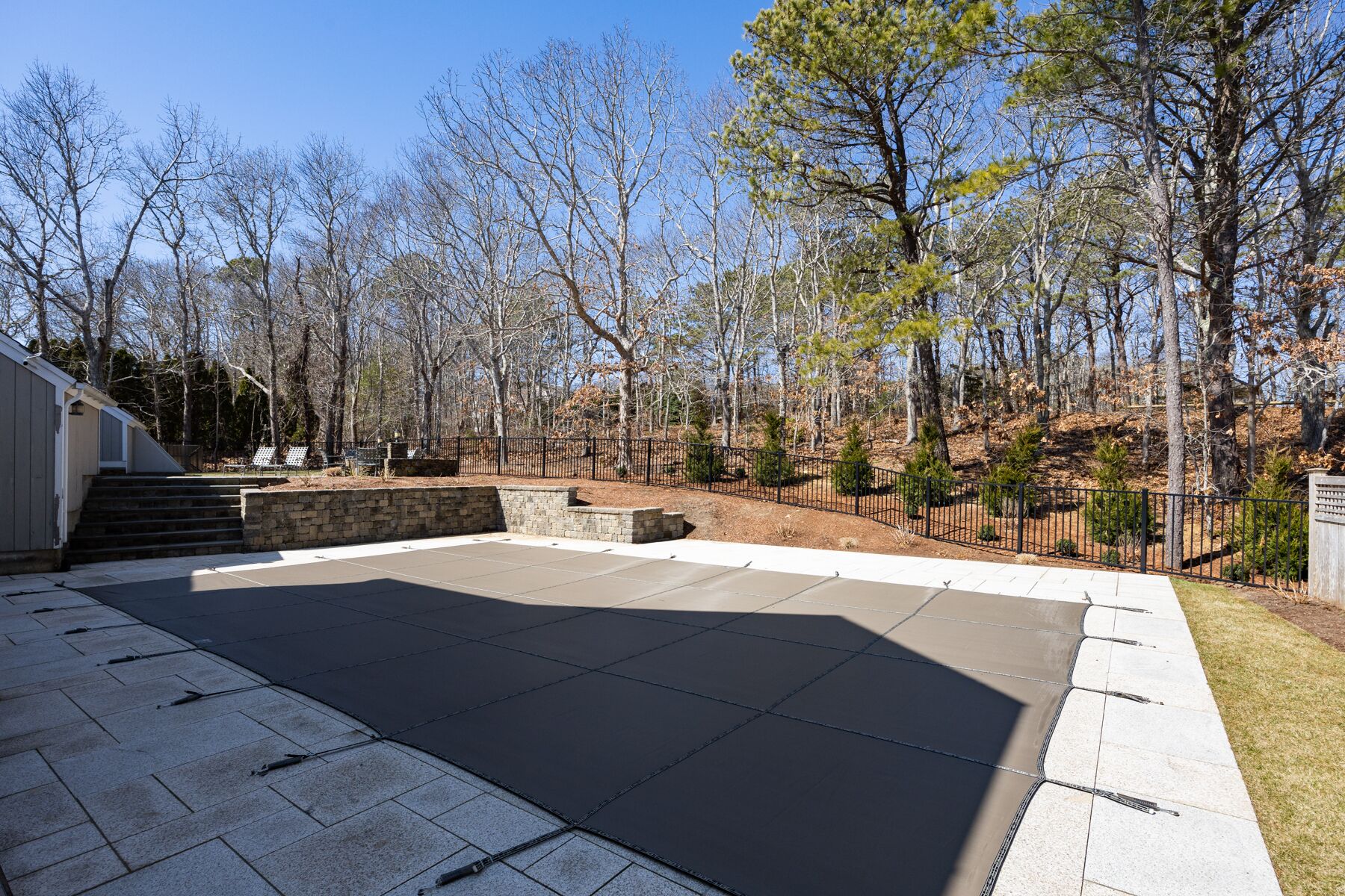 A backyard with a concrete patio, stone steps leading to a house, and a fenced area with trees and shrubs.