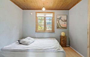 A bedroom with a wooden ceiling, a bed with white bedding, a window with blinds, a wall-mounted light fixture, and a framed picture. - Beach front home in Sjællands Odde (Sjællands Odde)