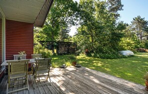 A wooden deck with a table and chairs, a covered outdoor area, and a garden with trees and a pile of firewood. - Beach front home in Sjællands Odde (Sjællands Odde)