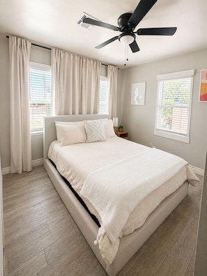 Camera da letto con un letto, ventilatore a soffitto, finestre con tende e un comodino con lampada.