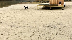 Pieni puinen mökki, jossa on pieni kuisti ja penkki, sijaitsee hiekkarannalla, ja lähellä on koira.
