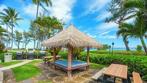 A thatched-roof gazebo with a blue cushion, surrounded by a stone pathway, palm trees, and a view of the ocean. - Updated Islander Studio | Ground Floor Convenience (Kapaa)