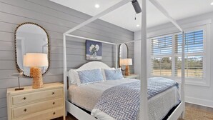 A bedroom with a canopy bed, two bedside lamps, a dresser, and a large window with blinds.