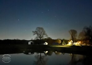 Eine nächtliche Szene mit einem Haus, das sich in ruhigem Wasser spiegelt, bewölktem Himmel und vereinzelten Sternen.