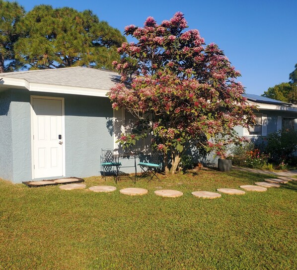 A house with a front yard, a lawn, a patio with chairs, and a tree with pink flowers.