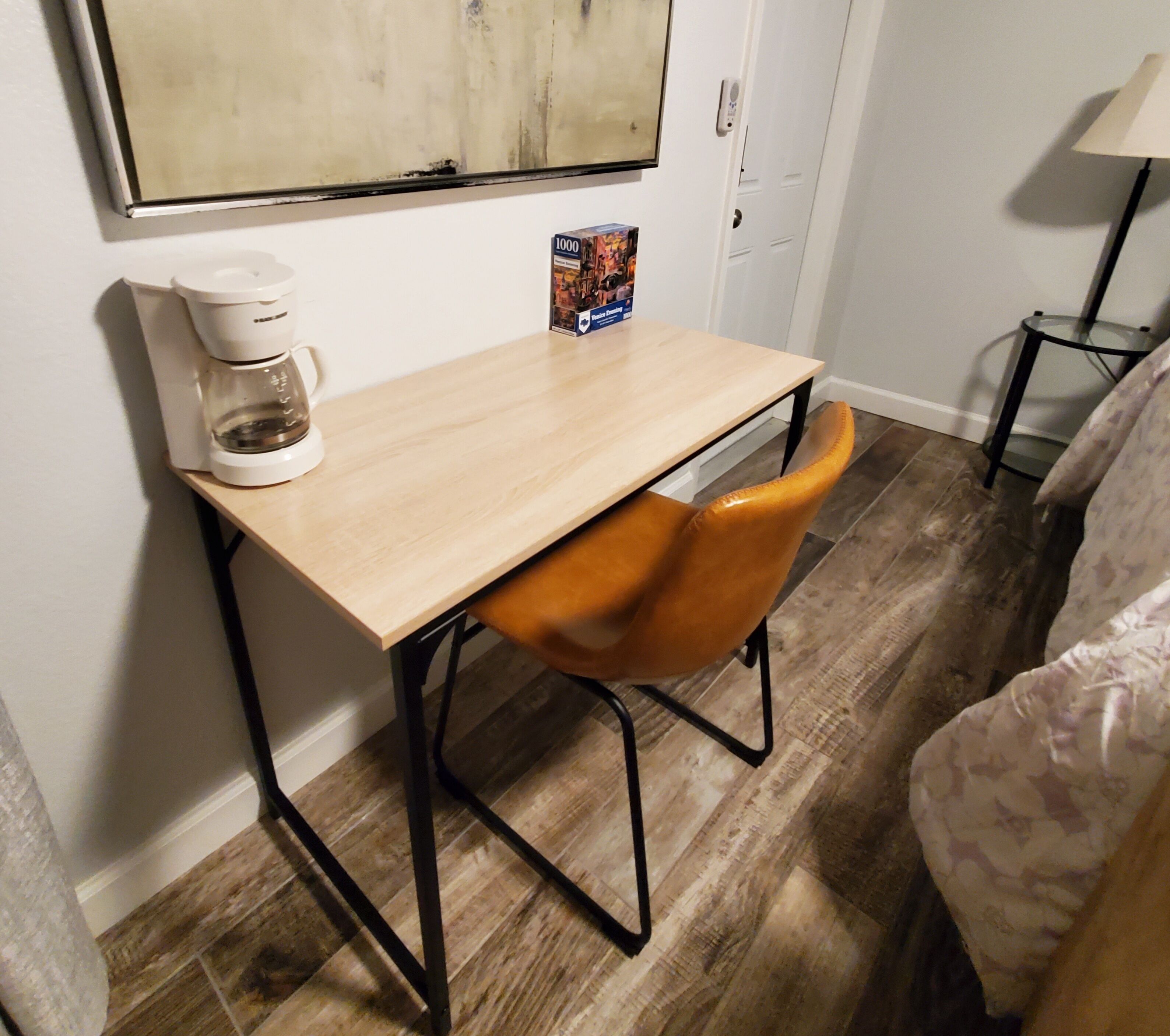 A small room with a wooden table, a chair, a coffee maker, and a framed picture on the wall.