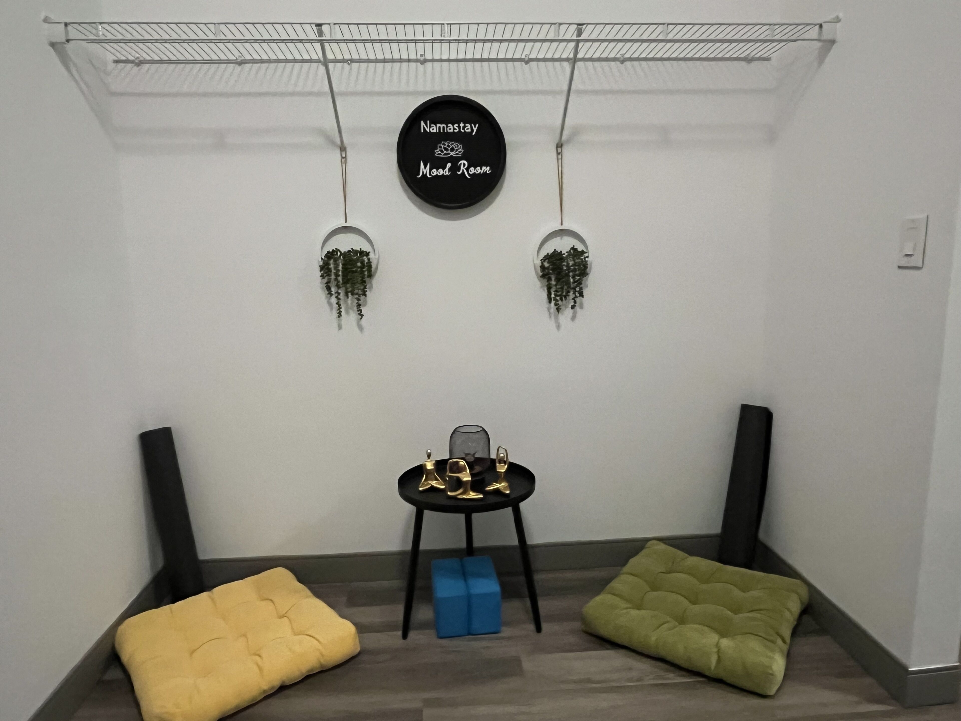 A room with a black circular sign reading "Namastay Mood Room," two hanging plants, a small table with a glass, and two colorful cushions.