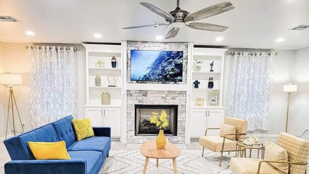 A living room with a blue sofa, beige armchairs, a fireplace, and a TV on the wall. - Hot Tub- Rose District-Walk to Shopping & Dining (Broken Arrow)