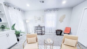A modern living room with two beige armchairs, a small round table, a white cabinet, and a dining table with chairs. - Hot Tub- Rose District-Walk to Shopping & Dining (Broken Arrow)